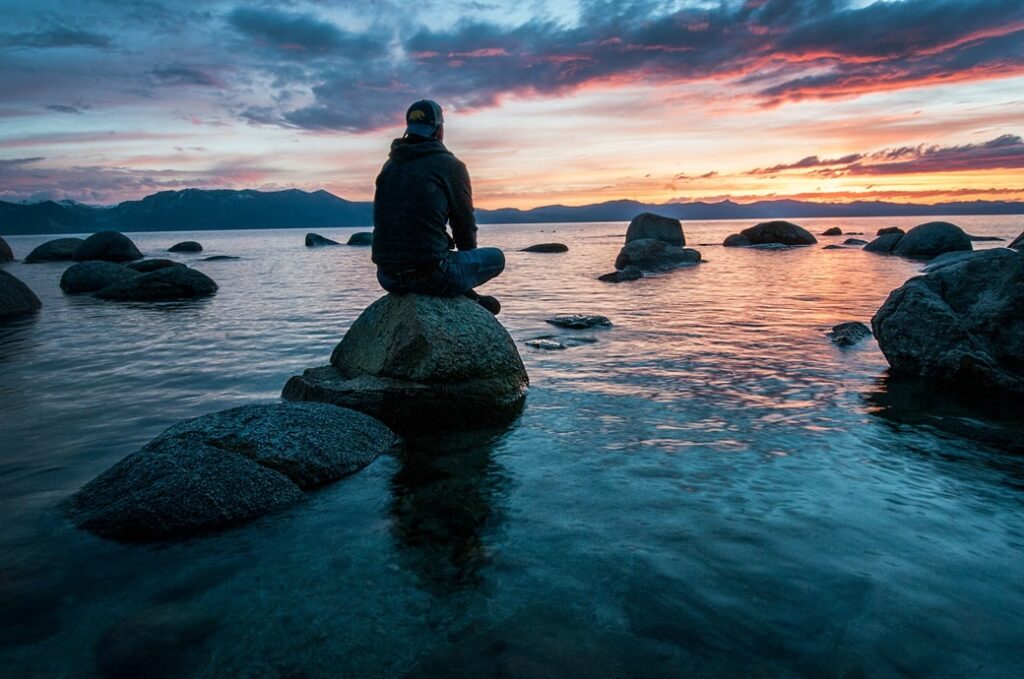 man sitting on rock in the ocean at sunrise setting goals
